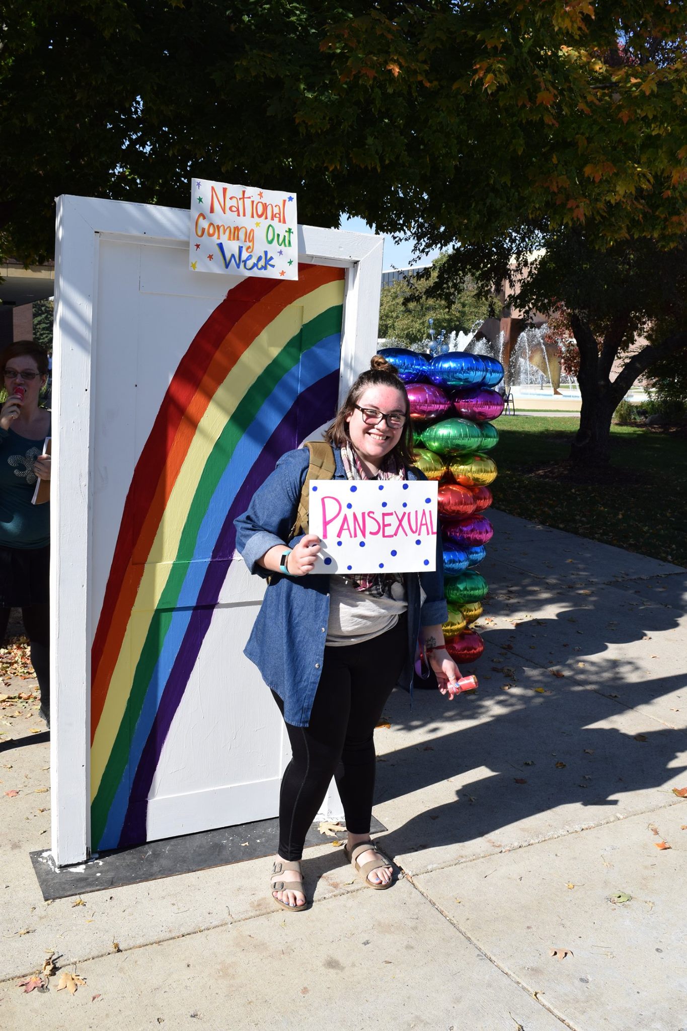 a person smiling outside of a rainbow-decorated door while holding a sign that says 'pansexual'