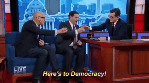 gif of Stephen Colbert toasting with two guests and saying 'here's to democracy!'