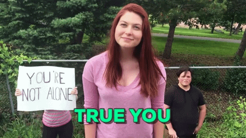 gif a woman smiling at the camera with two people in the background holding signs that 